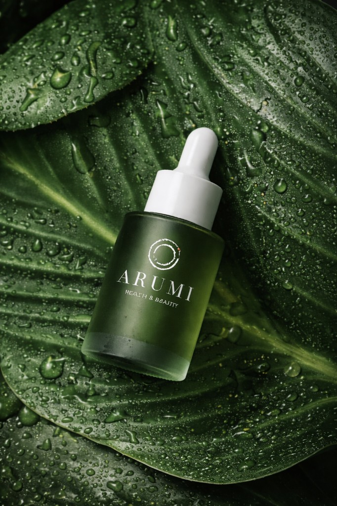 Arumi Health and Beauty — frosted green dropper bottle on dewy dark green leaves, white logo and serif wordmark